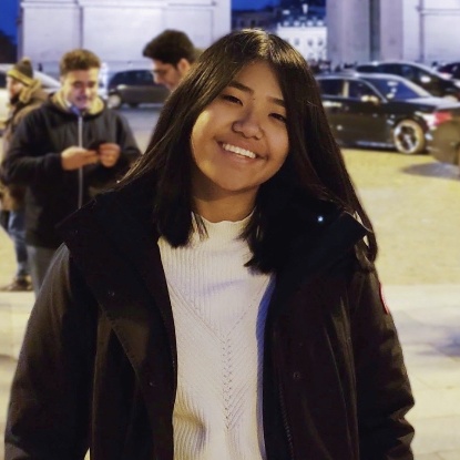 A smiling Asian person wearing a jacket standing outside in front of the Arc de Triomphe in Paris.