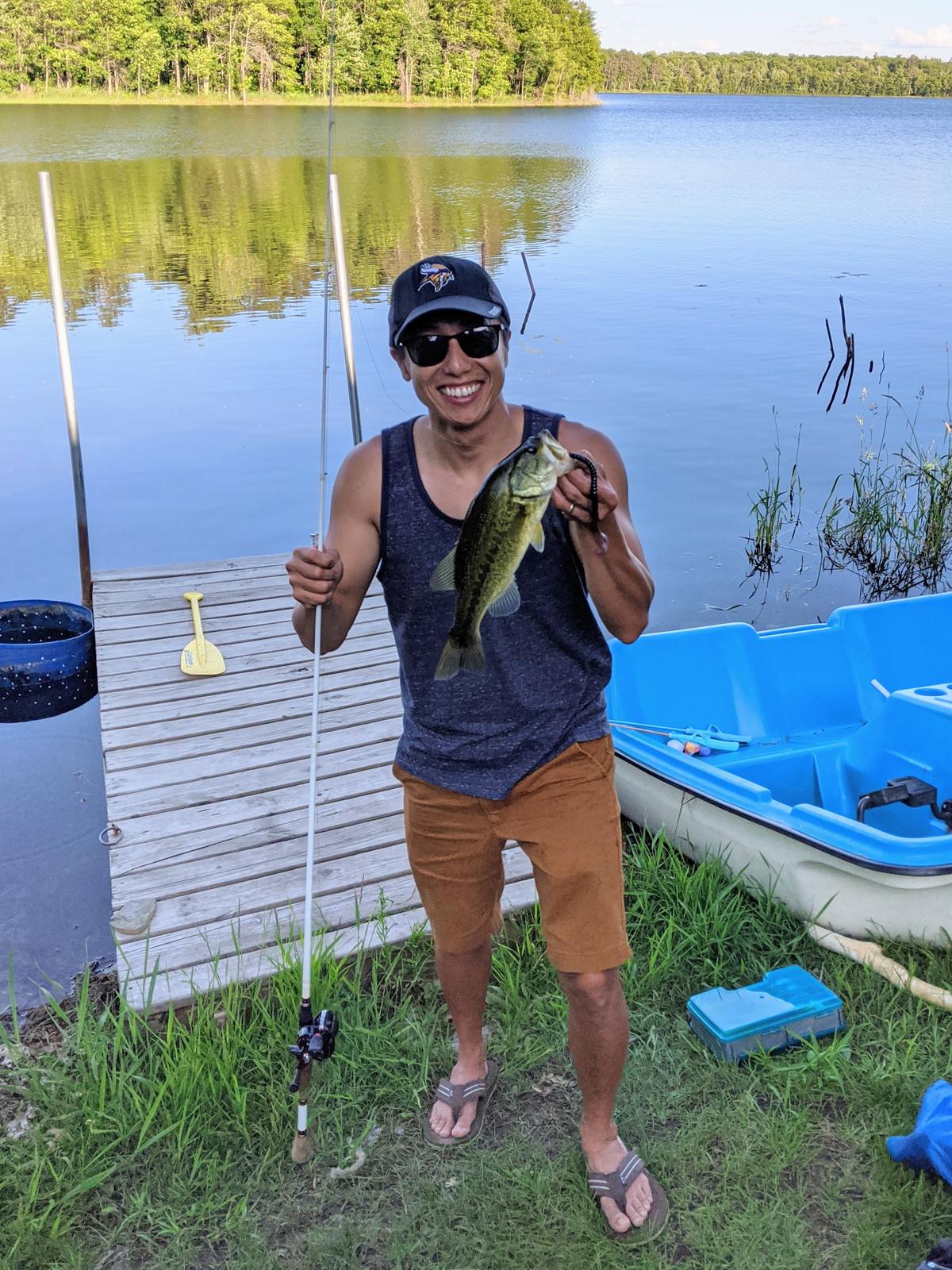 A smiling Asian man holding a fish and standing in front of a lake.