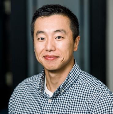 A smiling Asian man wearing a plaid check shirt. 