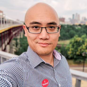 A selfie of a bald Asian man wearing glasses with an 'I voted' sticker on his shirt.